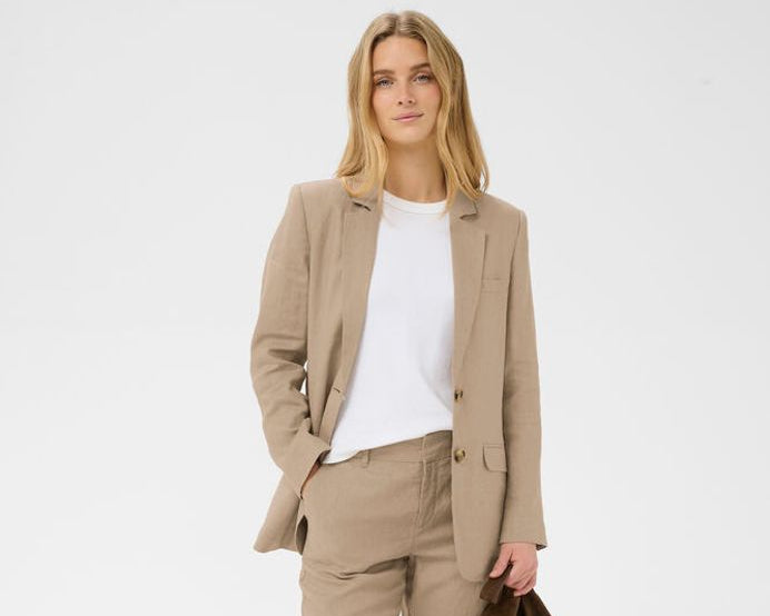Woman wearing a beige blazer, white shirt, beige pants, and brown sandals holding a brown bag on a white background